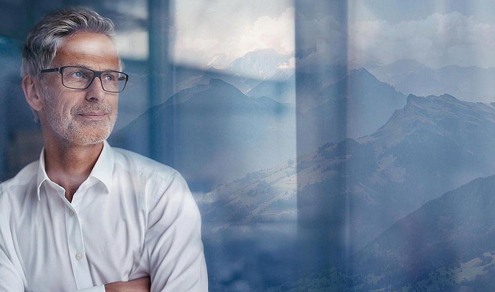 Ein Mann schaut mit einem leichten Schmunzeln durch eine Glasscheibe, im Hintergrund sind die Schweizer Berge zu sehen.