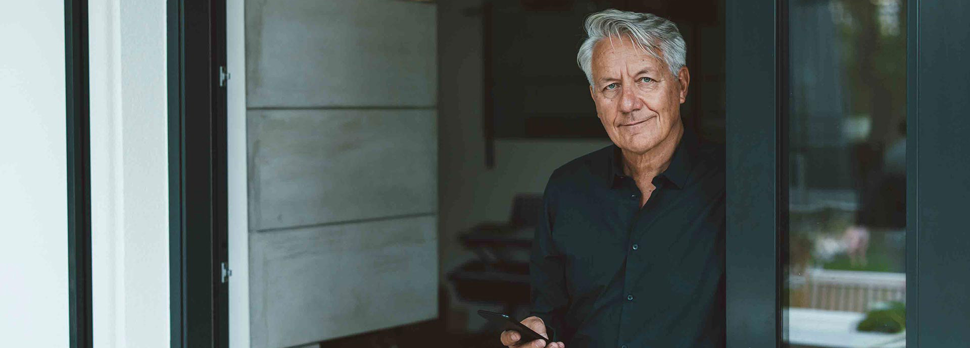 Ein Mann im Hemd steht im Glas-Türrahmen und hält sein Handy in der Hand