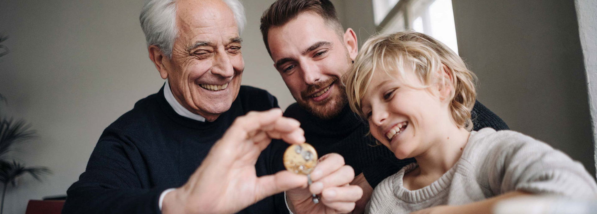 Drei Generationen, Großvater, Vater und Kind, lachen gemeinsam und schauen auf eine Münze, die der Großvater in der Hand hält.