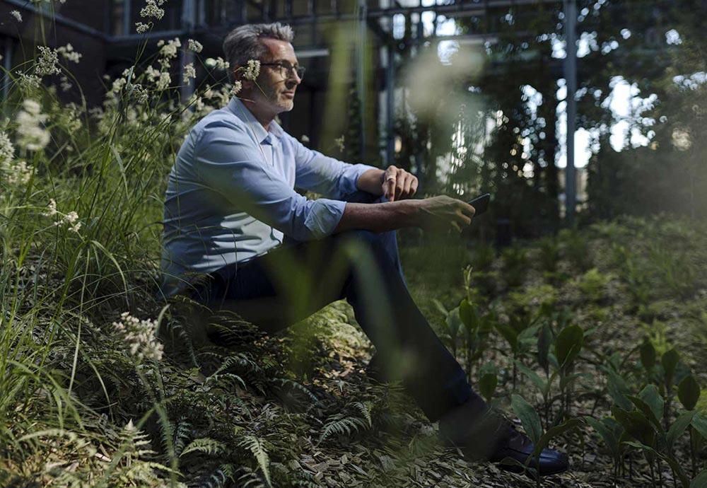 Ein Mann im Hemd sitzt auf der Wiese vor seinem Haus im Wald und hält sein Smartphone in der Hand.