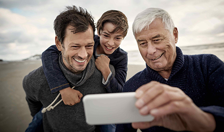 Ein Junge sitzt auf dem Rücken seines Vaters, neben ihm steht der Großvater. Gemeinsam schauen sie auf ein Handy und lachen.