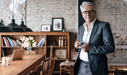 Ein Mann im grauen Anzug schaut schmunzelnd in die Kamera und hält eine Espresso-Tasse in der Hand.