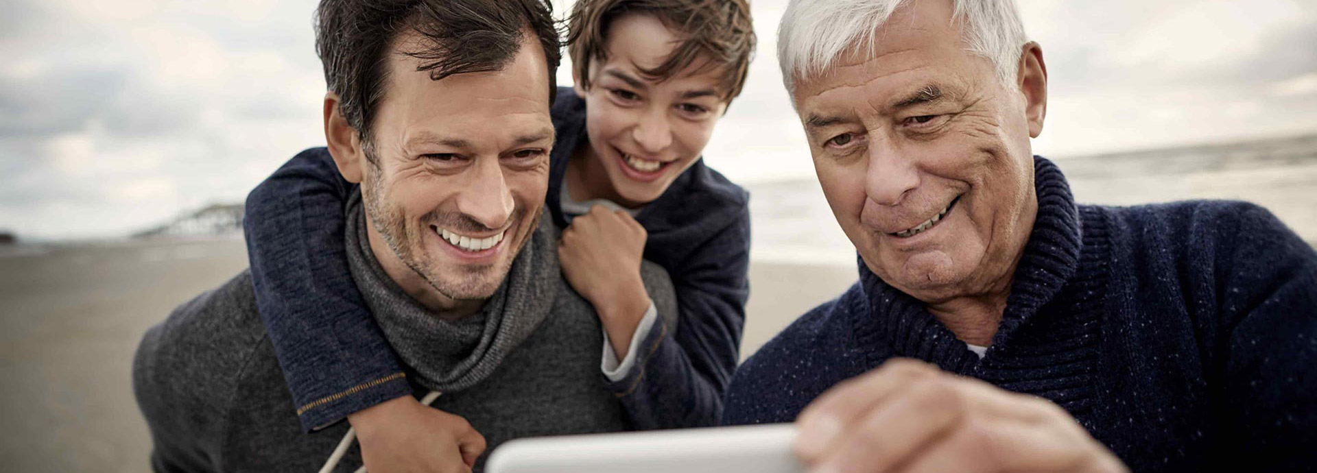 Ein Junge sitzt auf dem Rücken seines Vaters, neben ihm steht der Großvater. Gemeinsam schauen sie auf ein Handy und lachen.