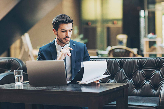 Ein Mann im Anzug sitzt auf einer Couch, mit einem Laptop vor sich. In seiner Hand hält er ein Notizbrett und blickt konzentriert darauf.