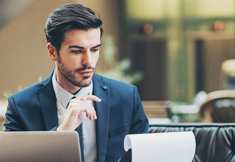 Ein Mann im Anzug sitzt auf einer Couch, mit einem Laptop vor sich. In seiner Hand hält er ein Notizbrett und blickt konzentriert darauf.