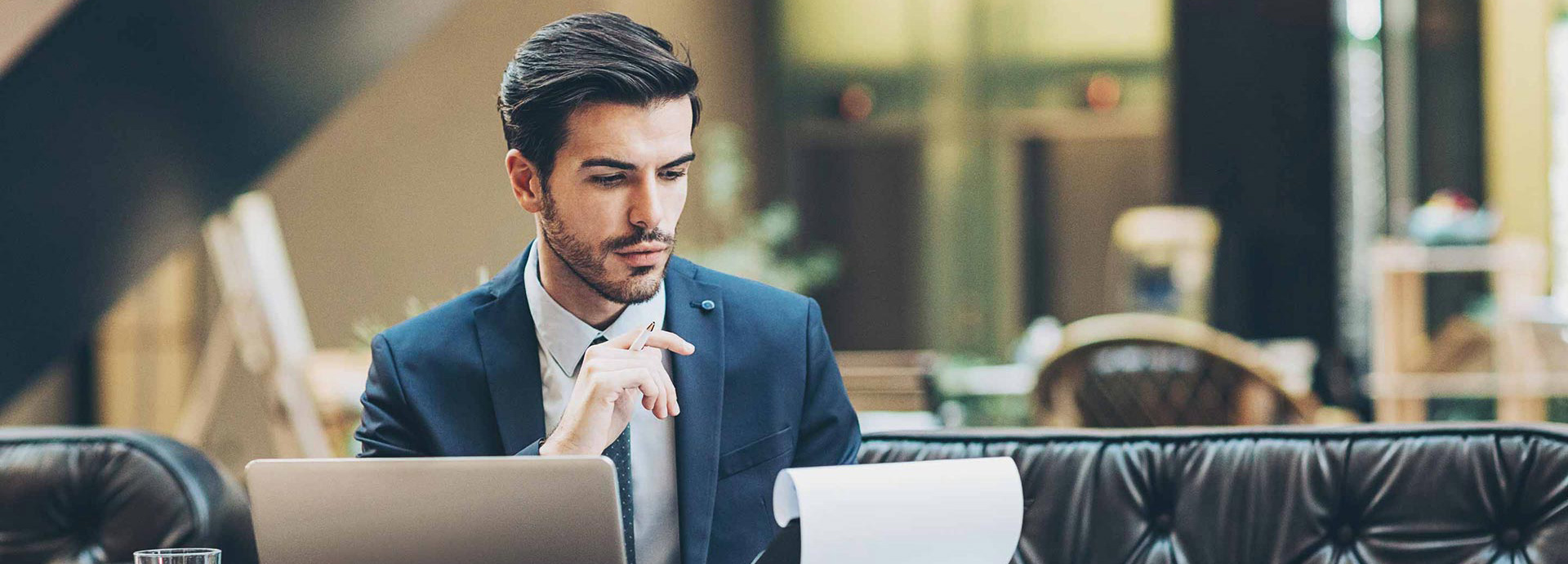 Ein Mann im Anzug sitzt auf einer Couch, mit einem Laptop vor sich. In seiner Hand hält er ein Notizbrett und blickt konzentriert darauf.