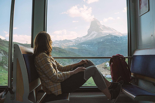 Frau schaut aus dem Fenster eines Zuges und sieht die Aussicht auf die Schweizer Berglandschaft mit grünen Hügeln und schneebedeckten Gipfeln im Hintergrund.