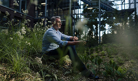 Ein Mann im Hemd sitzt auf der Wiese vor seinem Haus im Wald und hält sein Smartphone in der Hand.