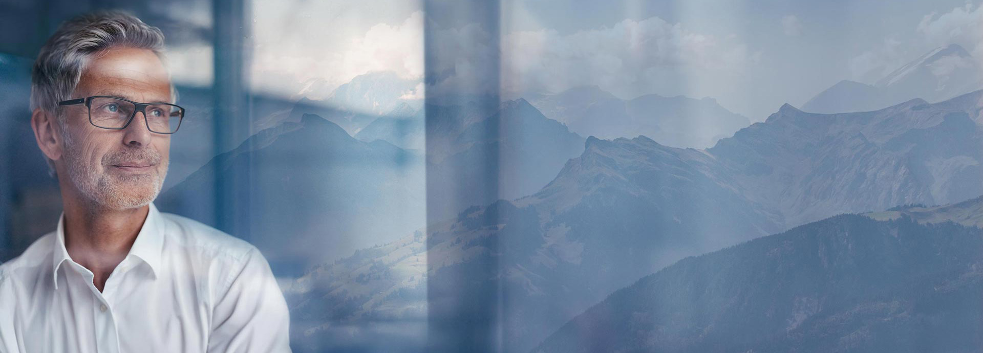Ein Mann schaut mit einem leichten Schmunzeln durch eine Glasscheibe, im Hintergrund sind die Schweizer Berge zu sehen.